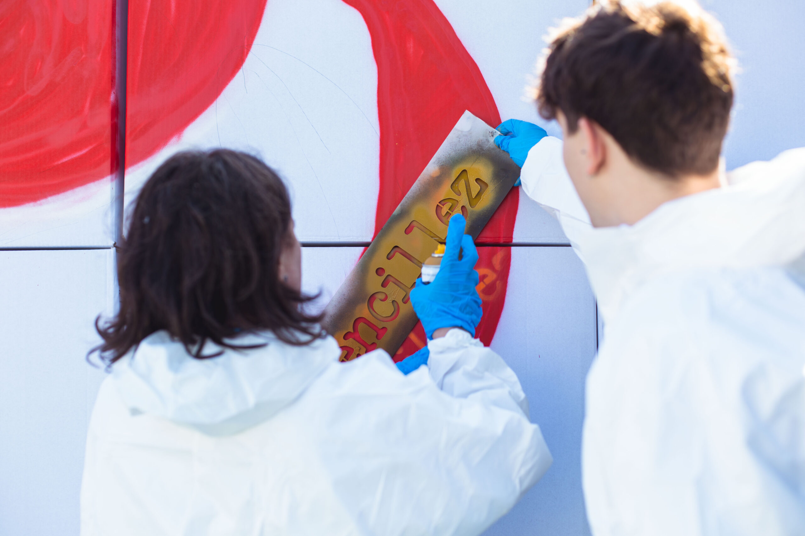 Dos personas con monos blancos y guantes azules pintando un mural con spray, mientras utilizan una plantilla para escribir 'pencil' sobre una pared blanca con trazos rojos. Actividad artística durante un evento del 75 aniversario de Inibsa.