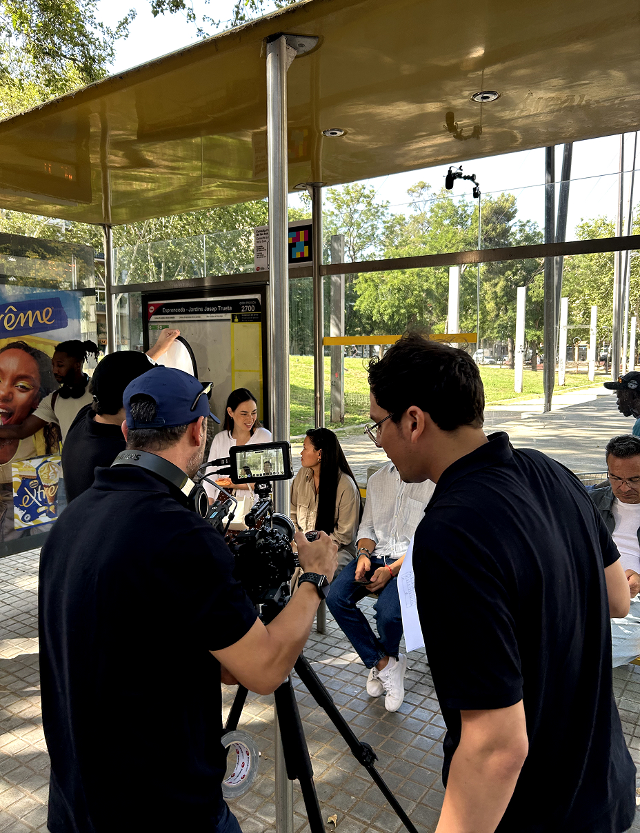 Grabación al aire libre en una parada de autobús, con un equipo de filmación capturando una escena, mientras las personas sentadas se preparan para la grabación.
